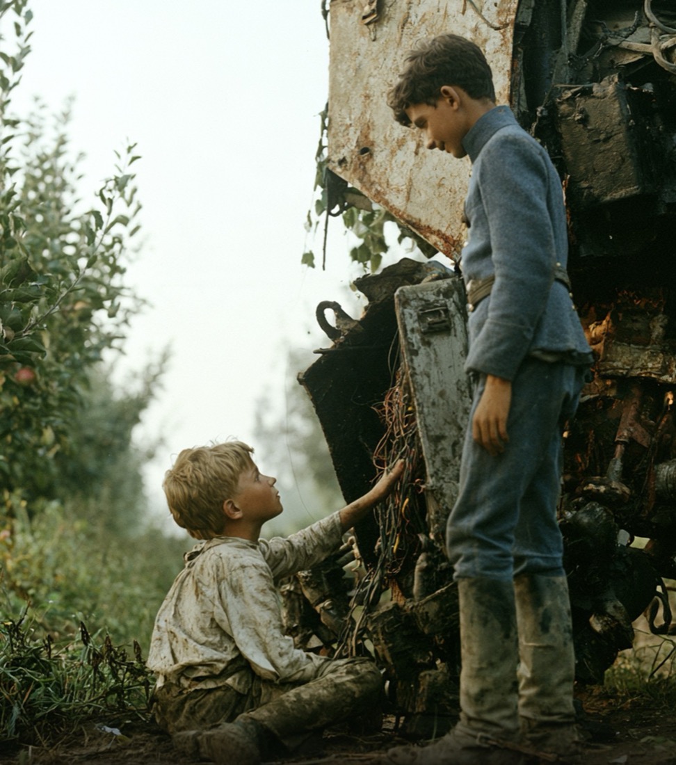 In an orchard, a small fair-haired boy sits in the dirt beside a wrecked machine, reaching into its wiring and looking up at his older brother, who stands over him in a grey-blue uniform.